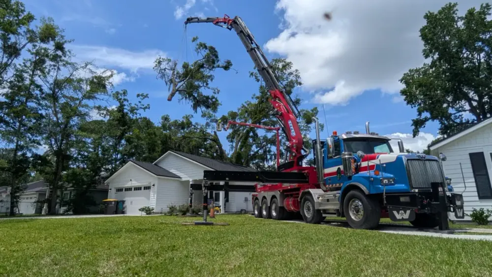 Jax Tree Removal blue Western Star truck with a red Effer knuckle-boom crane lifting a leafy oak limb over a driveway between two homes in Jacksonville, FL
