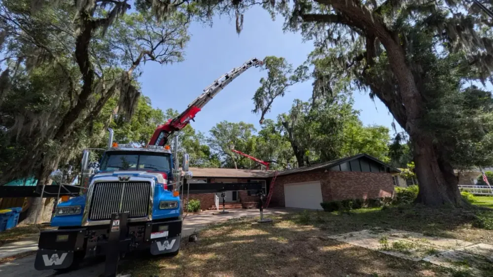 Crane boom arcing over a single-story brick home beneath a large live oak on a sunny day in Jacksonville, FL