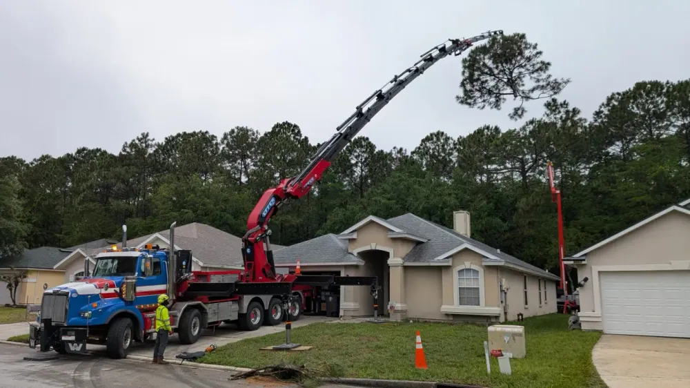 Blue crane truck with red knuckle-boom reaching to a tall pine over a stucco ranch home in Jacksonville, FL