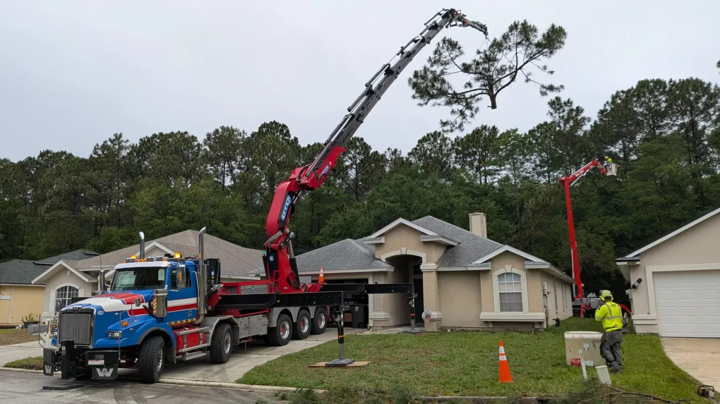 Jax Tree Removal crane boom and aerial lift working a tall pine above a stucco ranch home in Jacksonville, FL