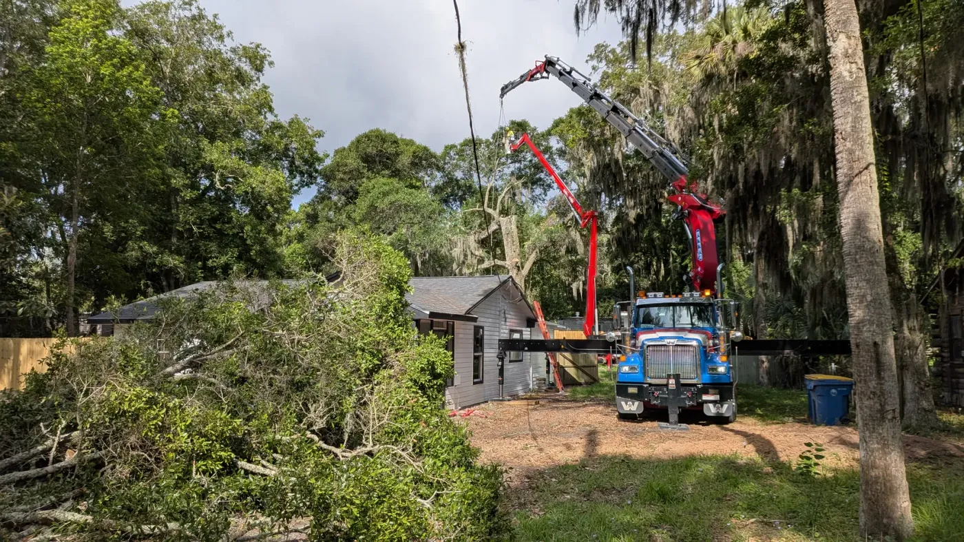 Jax Tree Removal crane truck reaching its boom over a small home with cut limbs on the ground during a backyard removal in Jacksonville, FL