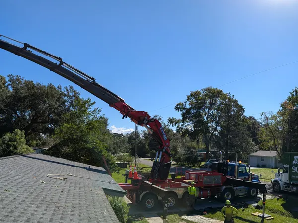 Panoramic view of a Jax Tree Removal knuckle-boom crane arcing over a rooftop to a topped tree, branded crane truck on site in Jacksonville, FL