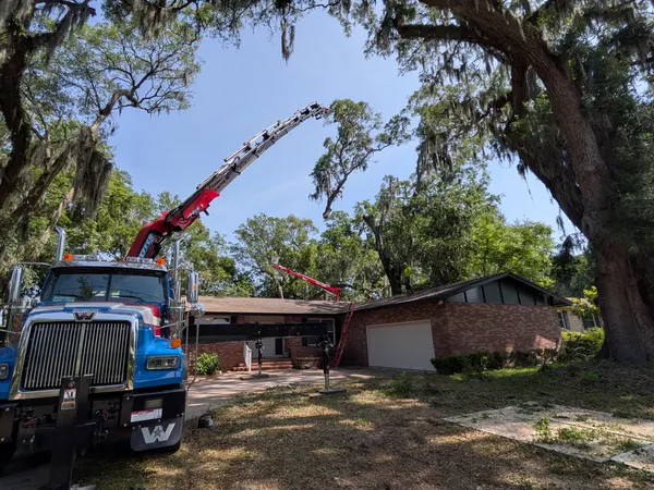 Crane boom arcing over a single-story brick home beneath a large live oak on a sunny day in Jacksonville, FL