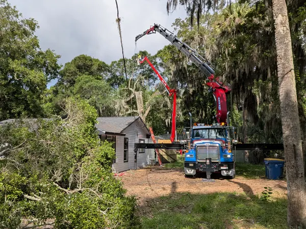 Jax Tree Removal crane truck reaching its boom over a small home with cut limbs on the ground during a backyard removal in Jacksonville, FL
