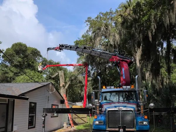 Front view of a Jax Tree Removal crane truck with boom and aerial lift extended over a home for a moss-draped oak removal in Jacksonville, FL