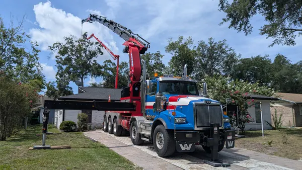 Blue Western Star knuckle-boom crane truck set up at a white brick ranch, a climber in the aerial lift topping an oak, crepe myrtles in bloom — crane tree removal in Jacksonville, FL by Jax Tree Removal